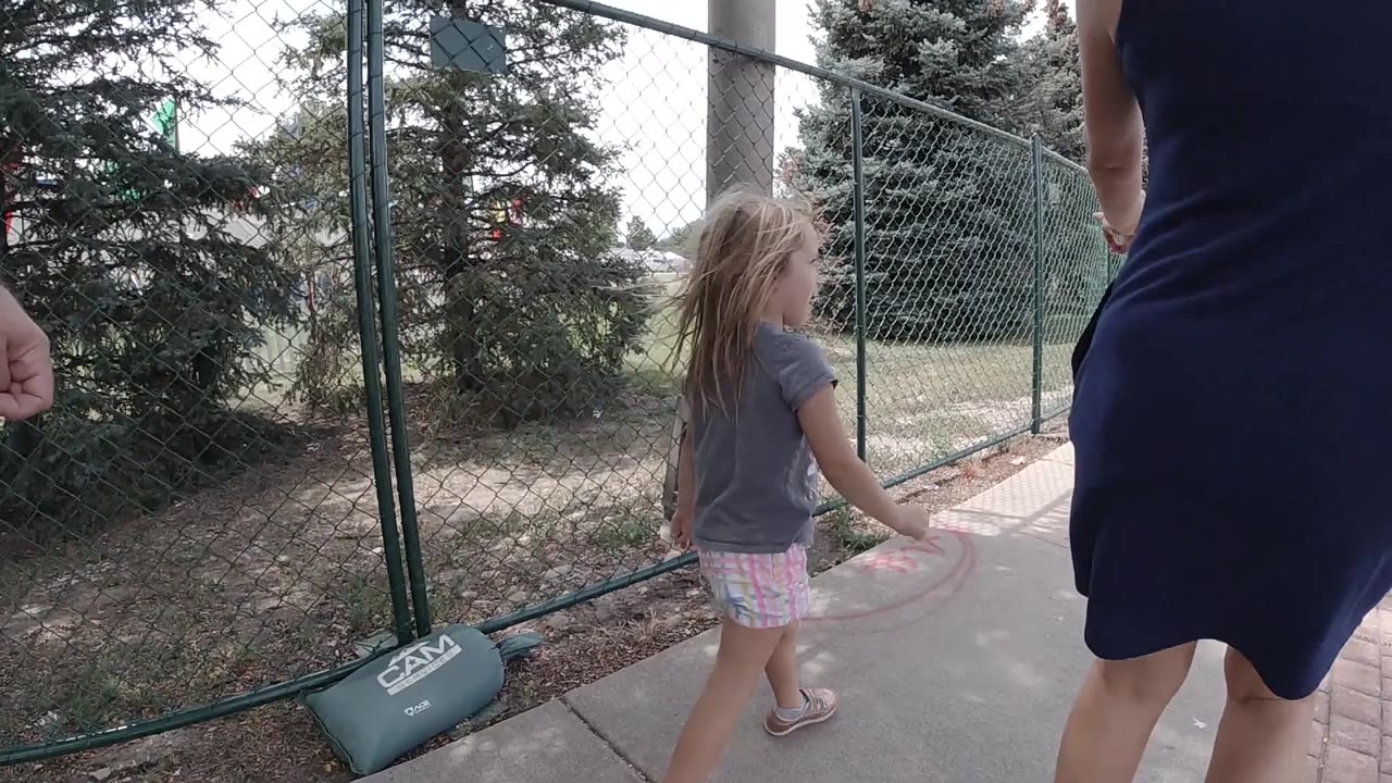 The girl continues forward on the sidewalk in Colorado while the adult remains cropped at the right edge and the chain-link fence runs beside the path.