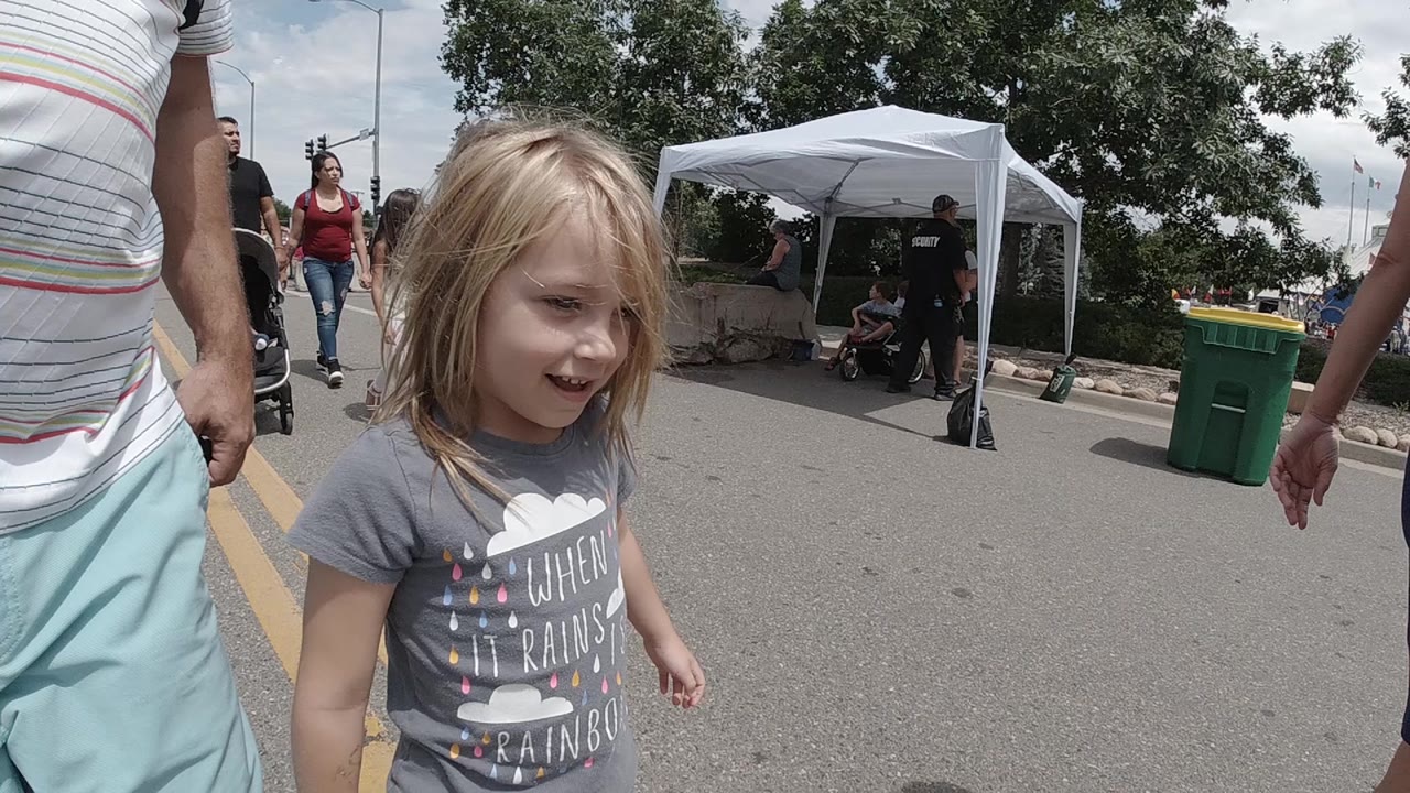 The girl remains near the foreground left on the Colorado street, looking ahead while adults, a stroller, white tent, trees, and green trash bins fill the background.