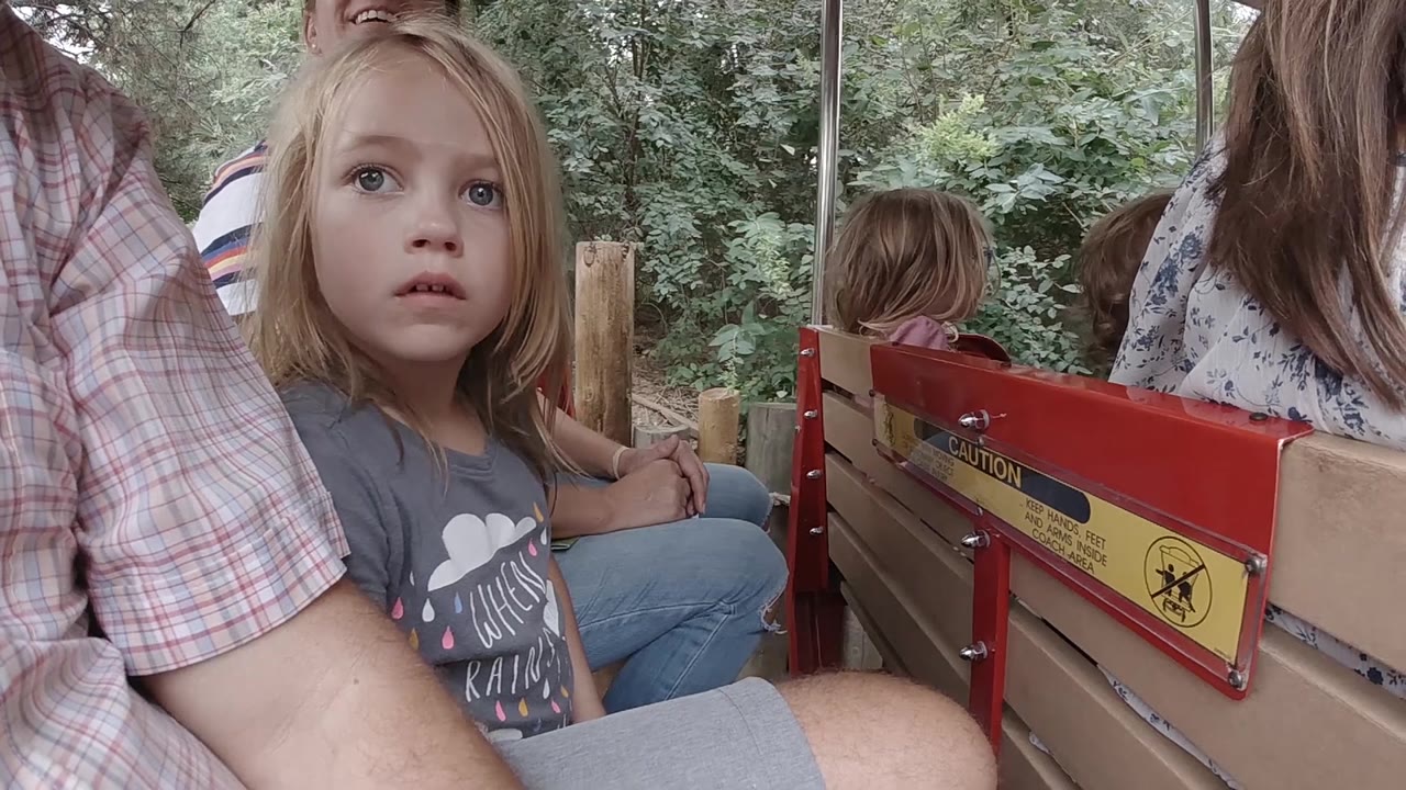 At 0:02, the girl remains seated near the camera as the red bench hardware and yellow warning label run across the middle of the Colorado zoo ride frame.