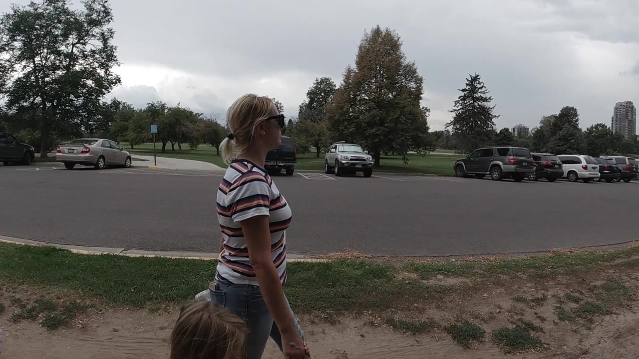 The woman and child remain in profile as they walk along the path, with a line of cars across the road and broad park grass behind them in Colorado.
