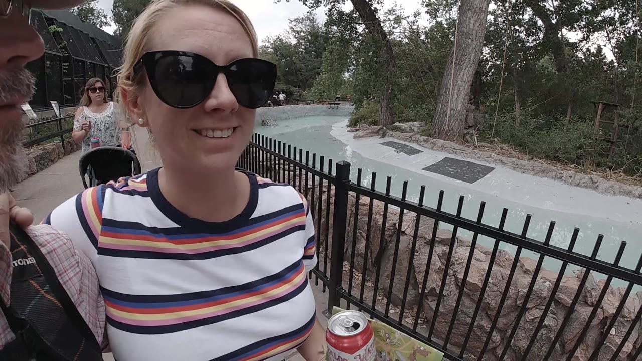 A close view shows the woman smiling at the camera, with the black metal fence, empty path curve, and wooded exhibit area visible over her shoulder in Colorado.