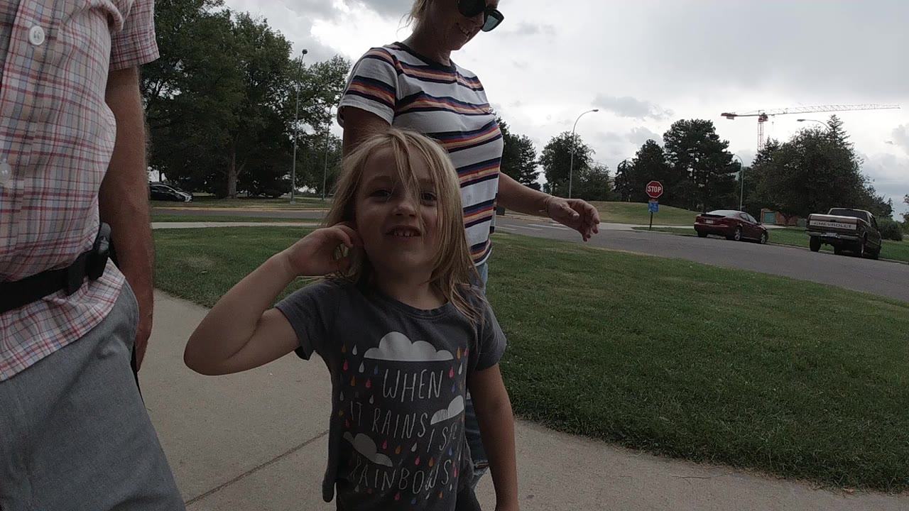 In Colorado, United States, a young blonde girl raises her hand near the camera while two adults are cropped beside her on a sidewalk by grass and a road.