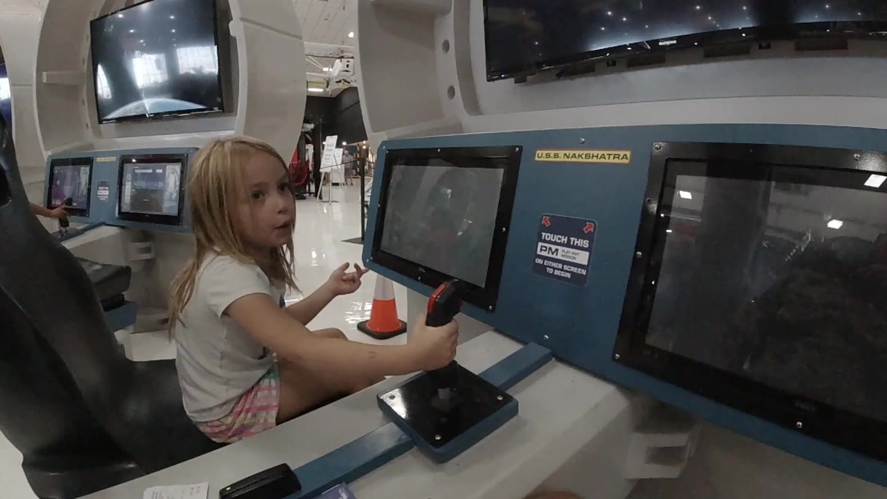 At 0:00, a young blonde girl sits beside a blue museum exhibit console in Colorado, Mexico, with her hand near a black joystick and dark display screens in front of her.