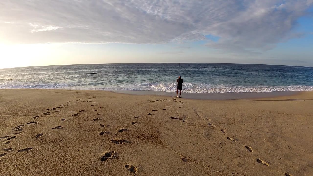 A wide Los Cabos shoreline view shows the person centered near the surf while footprints curve through the foreground sand and low waves roll in.