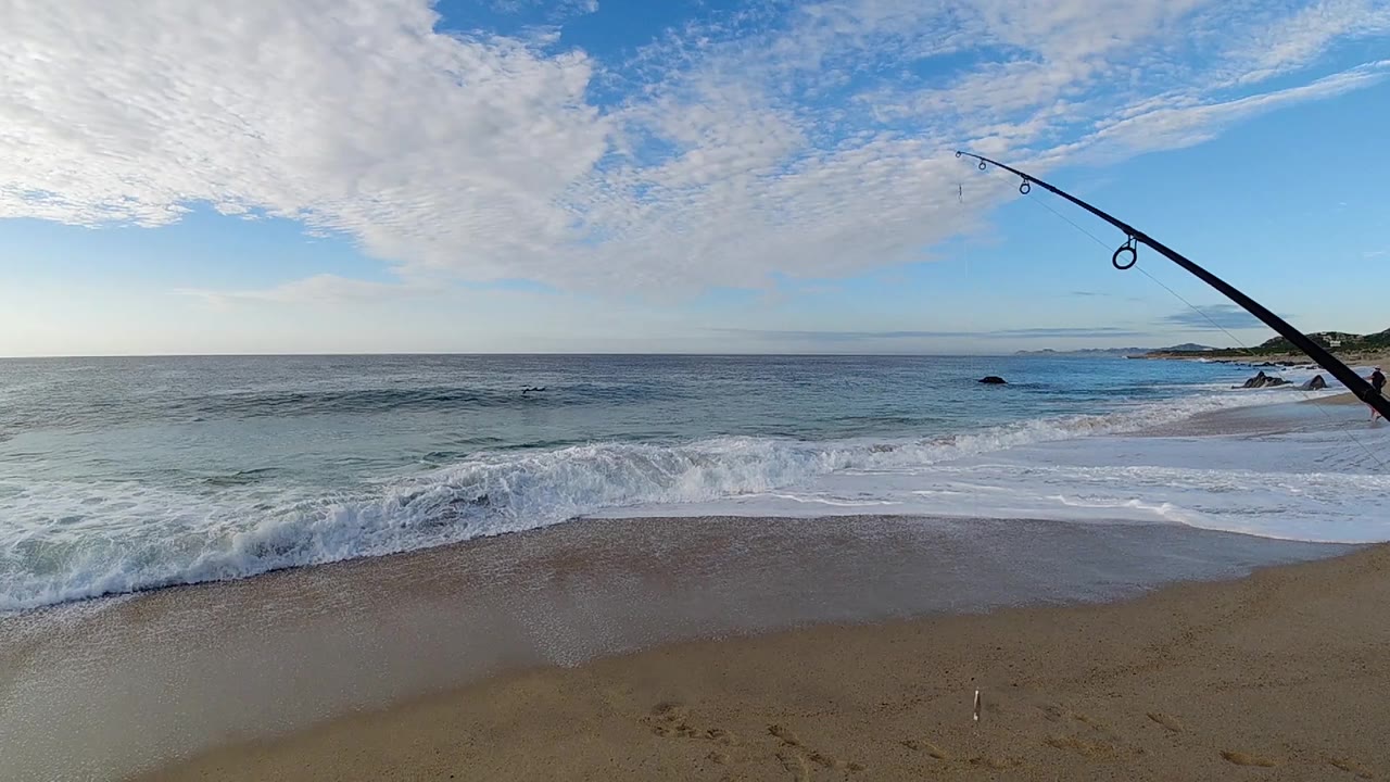 At the opening moment in Los Cabos, Mexico, a dark fishing rod angles in from the right above a sandy beach and low blue surf, with no recognizable people visible.