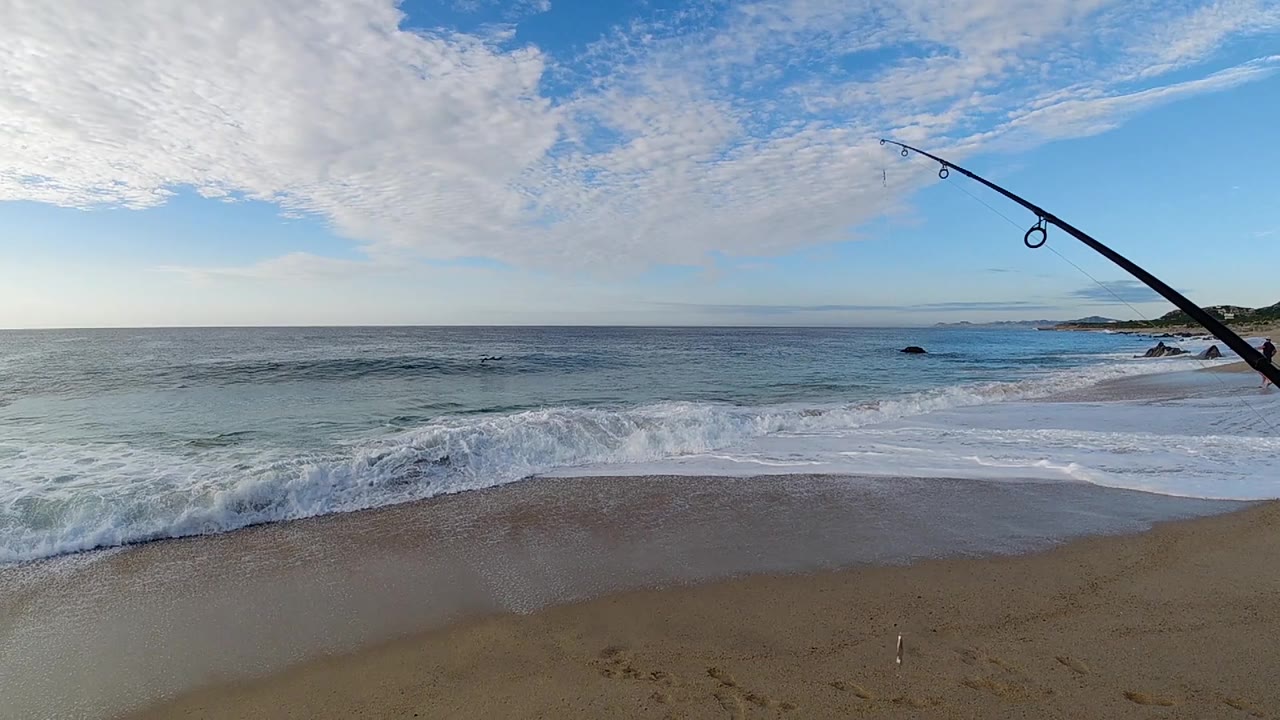 A thin wave line moves closer to the wet sand while the fishing rod remains at the right edge above the Los Cabos shoreline, with no recognizable people visible.