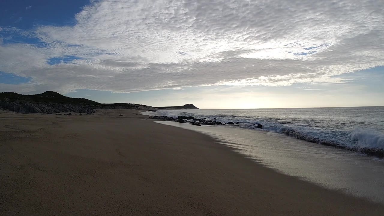 Opening frame of a Los Cabos beach with dark sand in the foreground, surf on the right, distant hills on the left, layered clouds overhead, and no visible people.