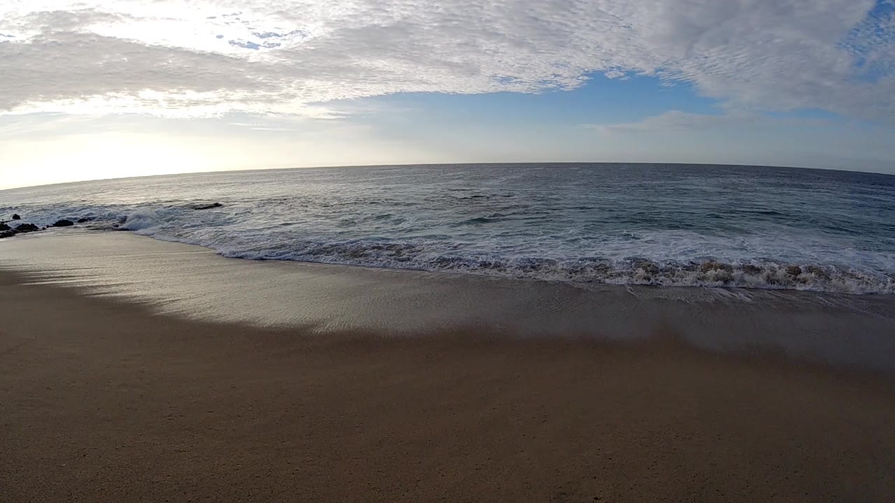 Opening view of the Los Cabos shoreline with damp sand in front, low surf at center, bright sun behind clouds to the left, and no visible people.