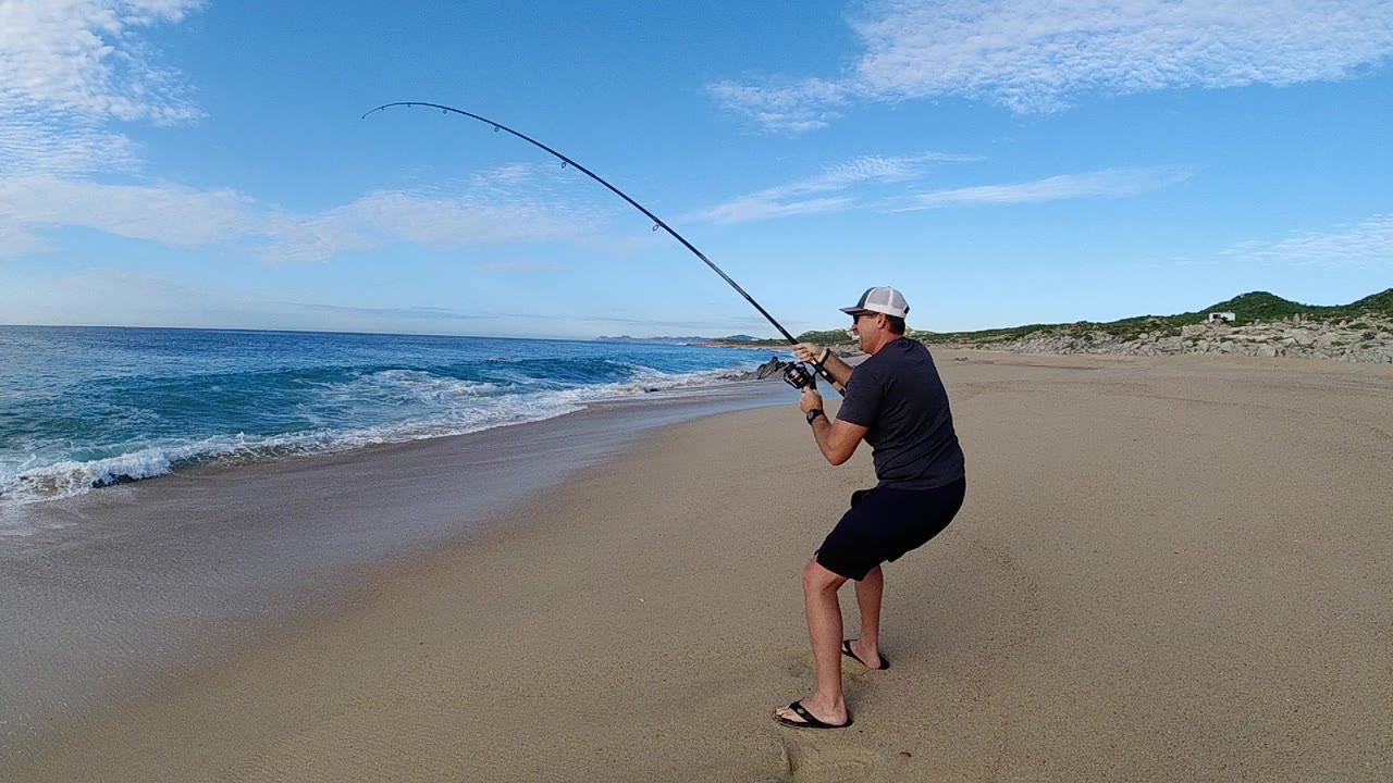 At 0:00, an adult man in a cap braces on the Los Cabos beach with a fishing rod bent over the blue surf, with open sand and low dunes behind him.