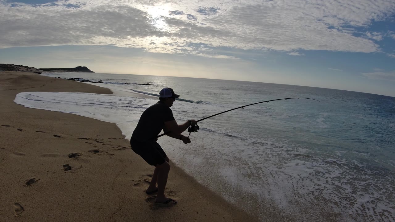 Silhouetted man fishing from a sandy beach beside gentle surf.
