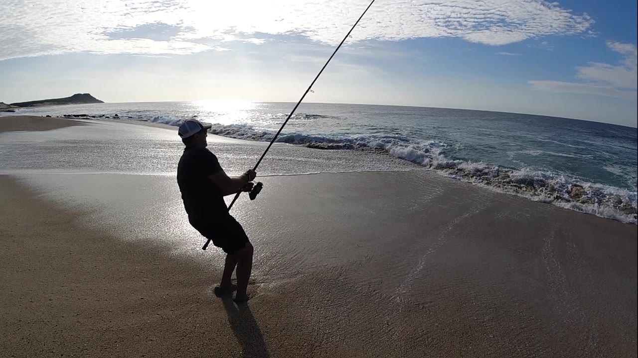 A backlit fisherman leans away from the surf in Los Cabos, Mexico, gripping the reel as foam washes across the beach behind him.