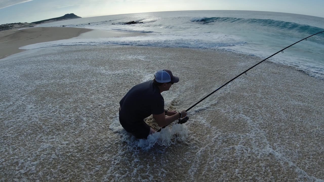 Man kneeling in shallow surf with a fishing rod on a sandy beach.