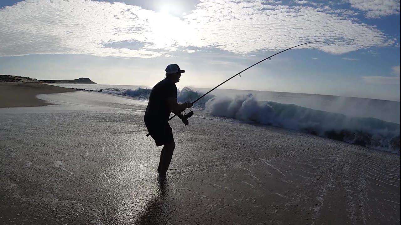 A lone fisherman braces on the wet beach while a low wall of surf moves in from the right and the rod extends across the sky.