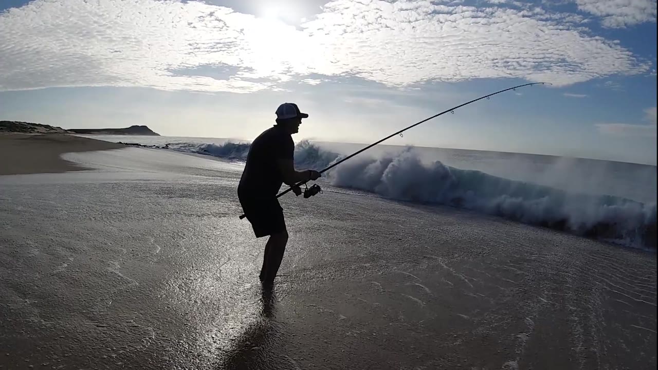 Fisherman lowers his stance beside a breaking wave.