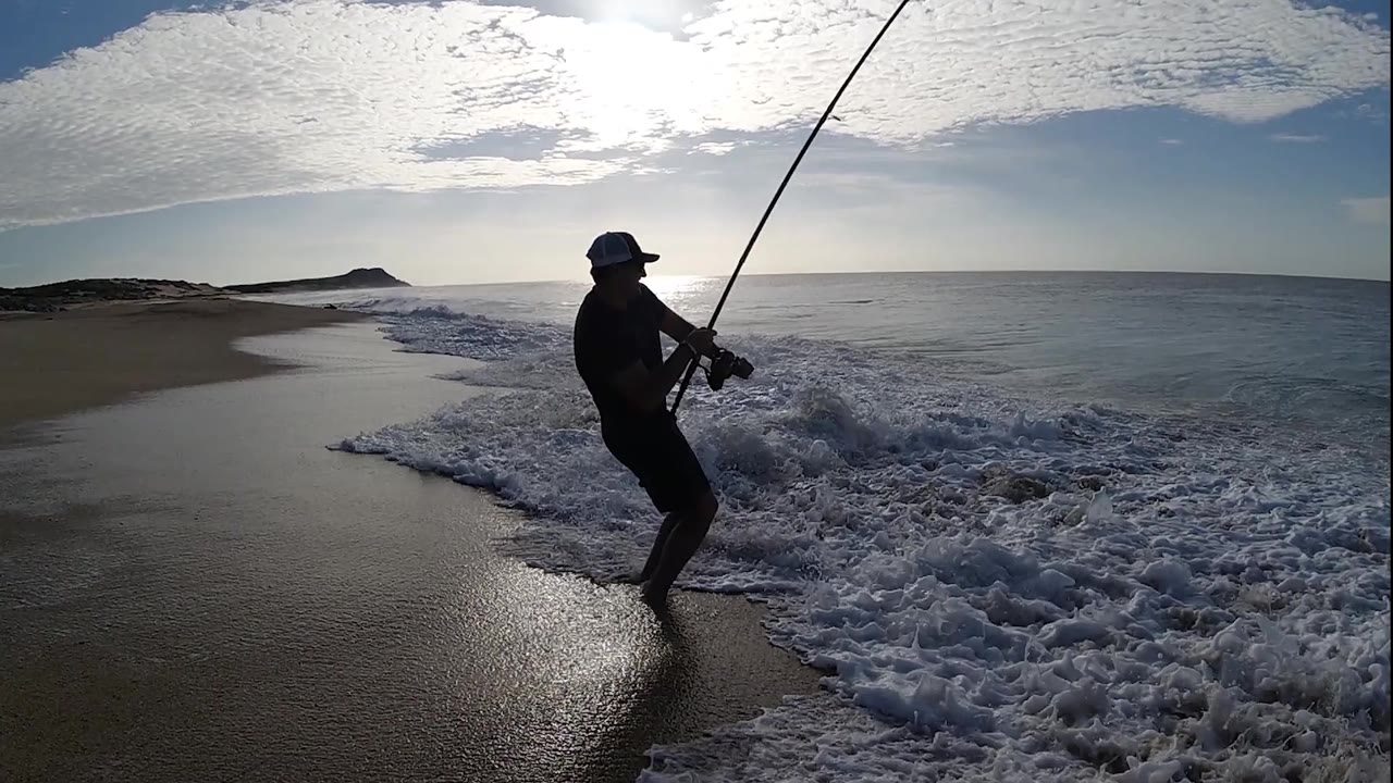 Fisherman braces near incoming whitewater with rod angled upward.