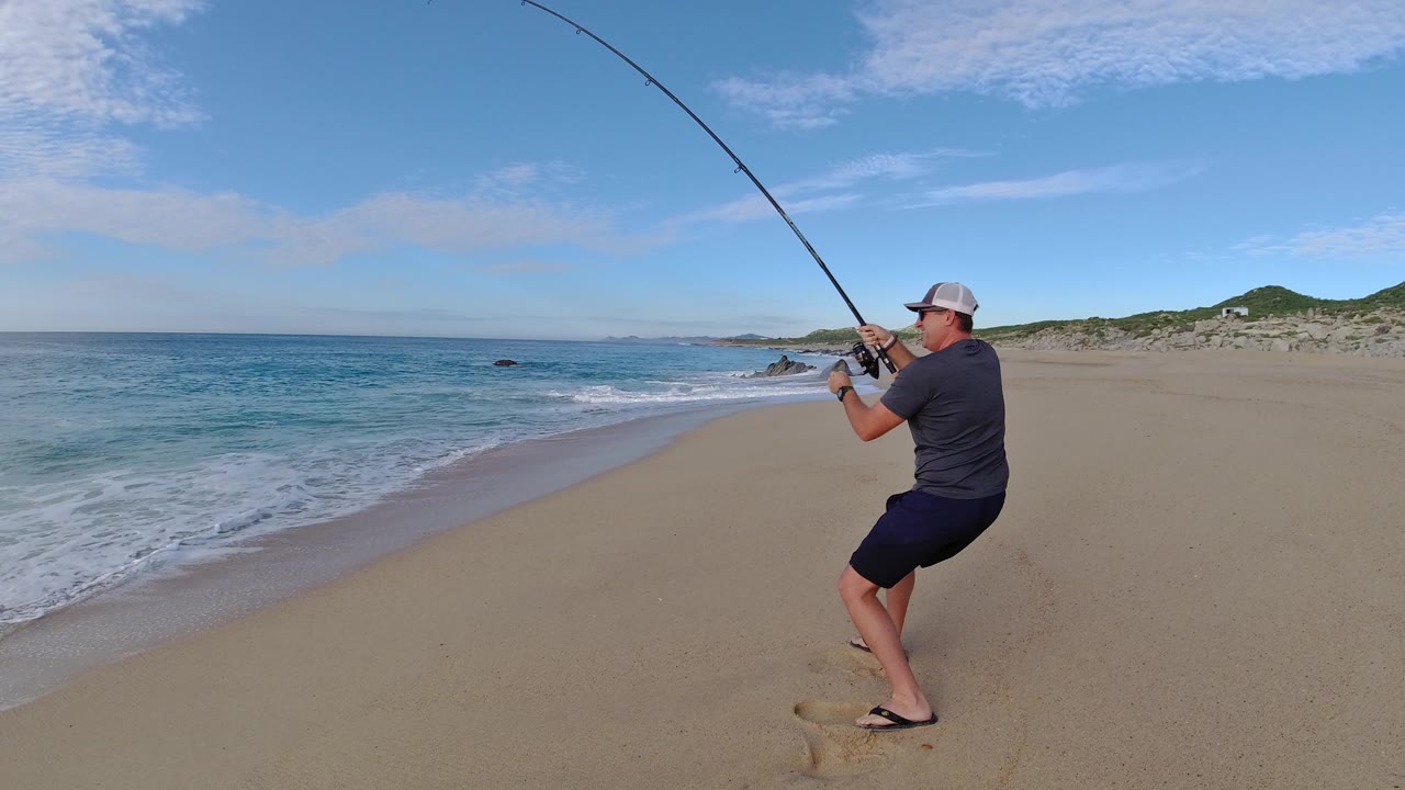Man surf fishing on a sandy beach with a bent rod.