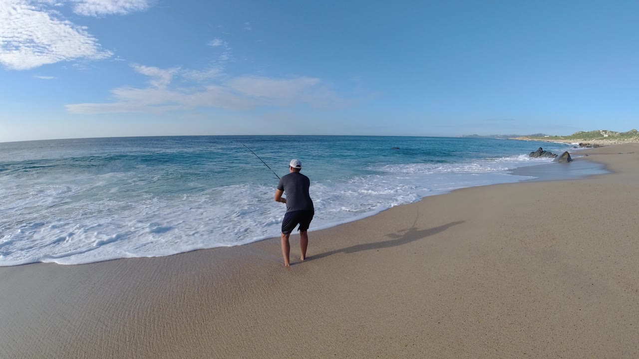 The fisherman remains centered on the Los Cabos beach while foamy water curls in from the left and the horizon stays level behind him.
