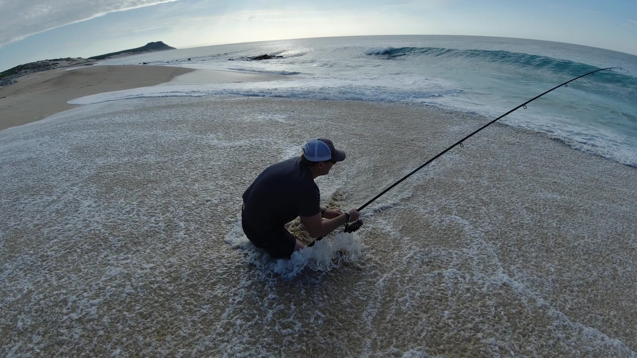 Man crouching in shallow surf with a fishing rod on a beach.