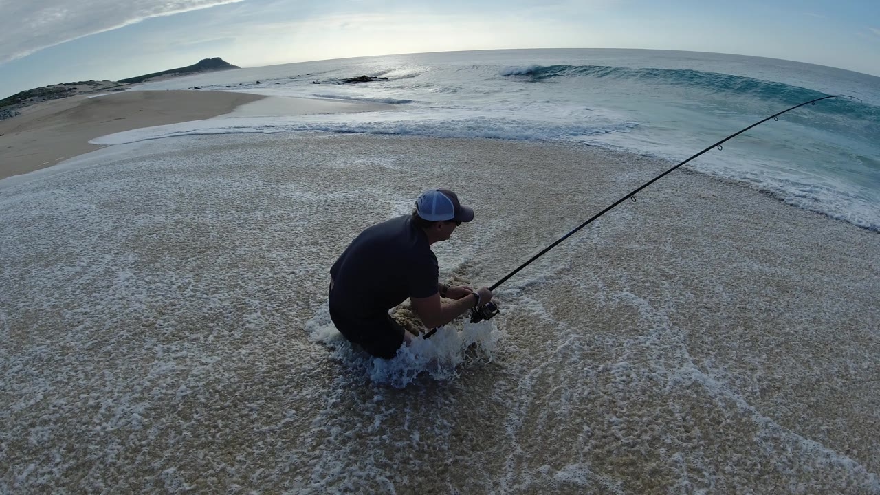 The fisherman remains crouched in shallow water at the Los Cabos shoreline, with the rod angled right across the frame and foam washing around his legs.