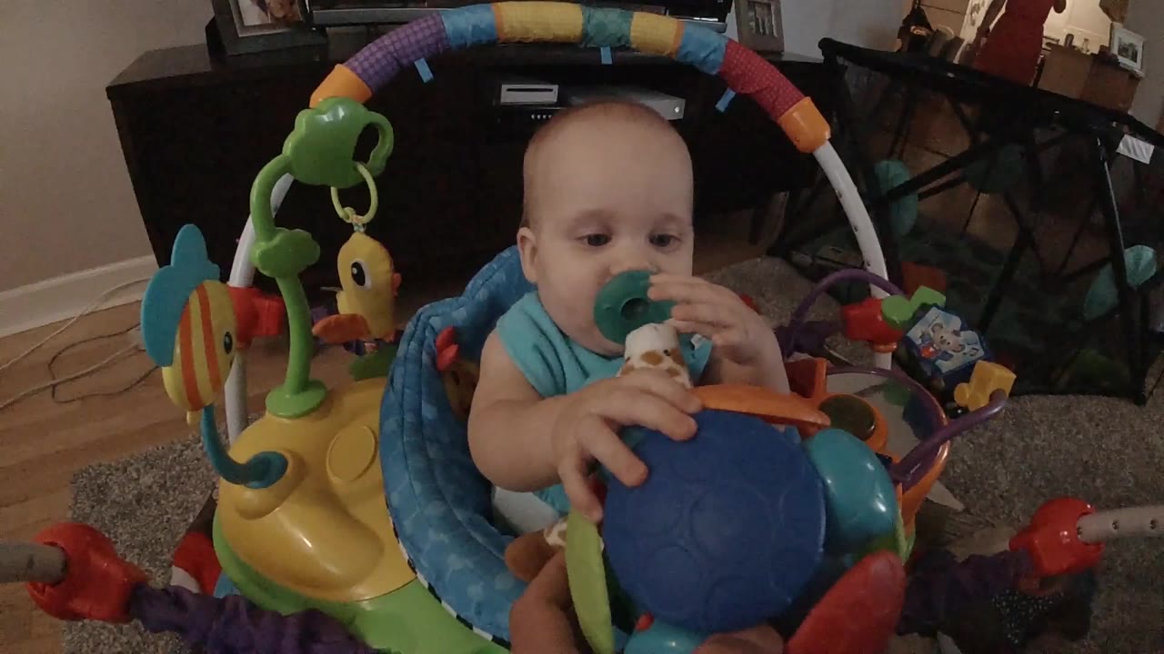 At 0:01, the baby boy looks toward the colorful ball in front of him, with hanging activity toys and dark furniture behind the seat.
