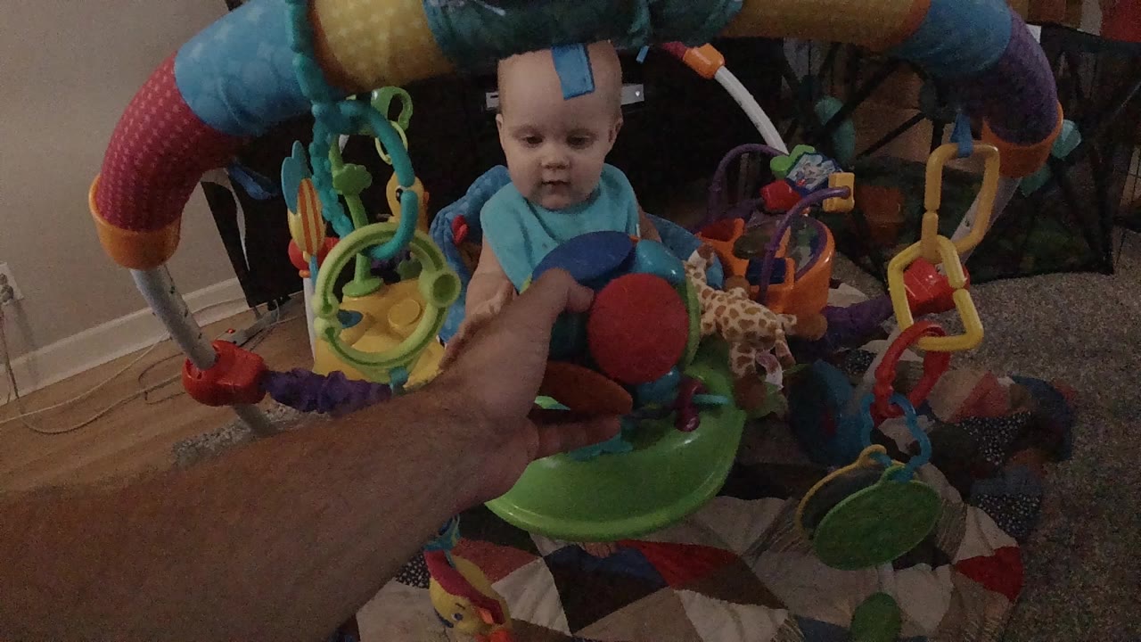 Baby in activity center facing a colorful ball held by an adult hand.