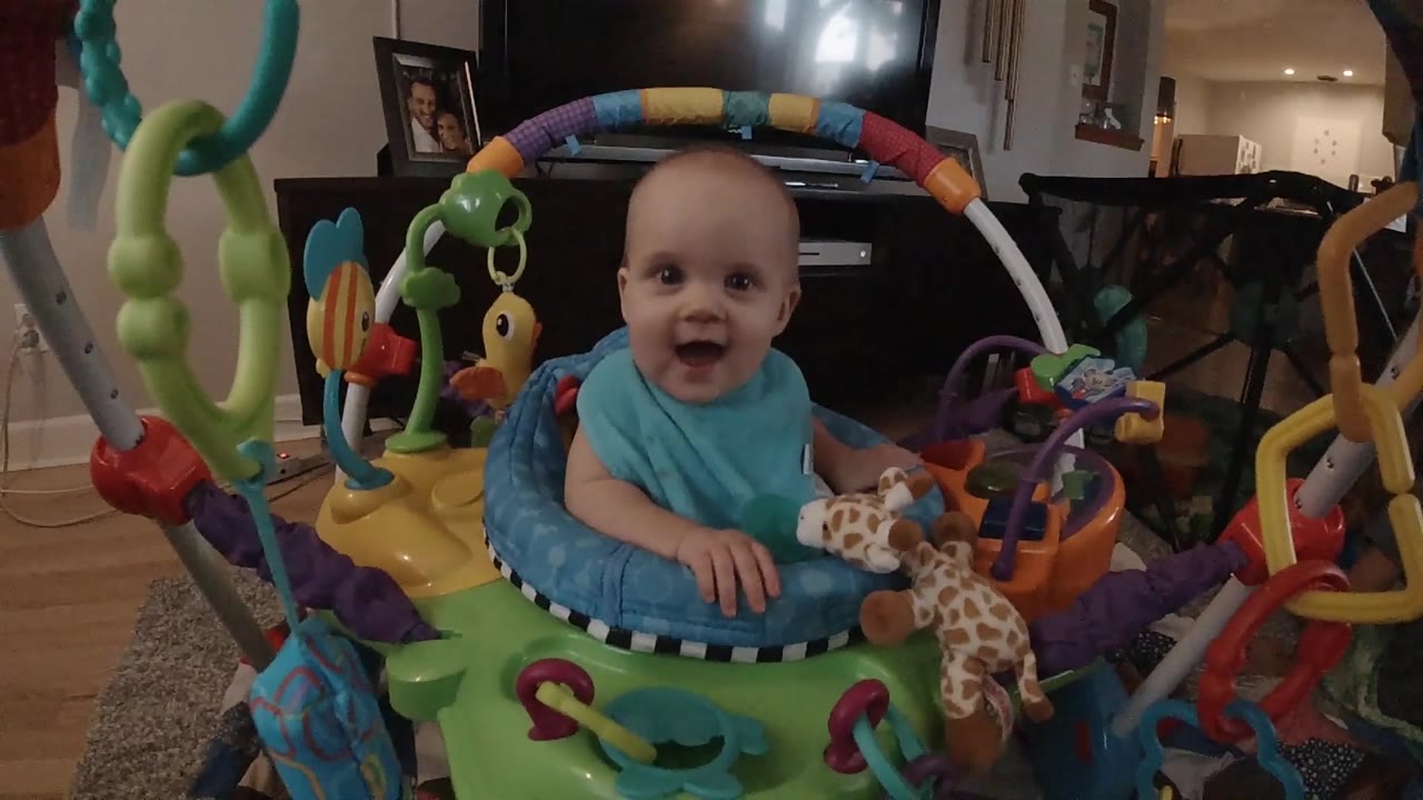 At 0:02, the baby faces the camera from the padded jumper seat with a plush giraffe hanging near the right hand.
