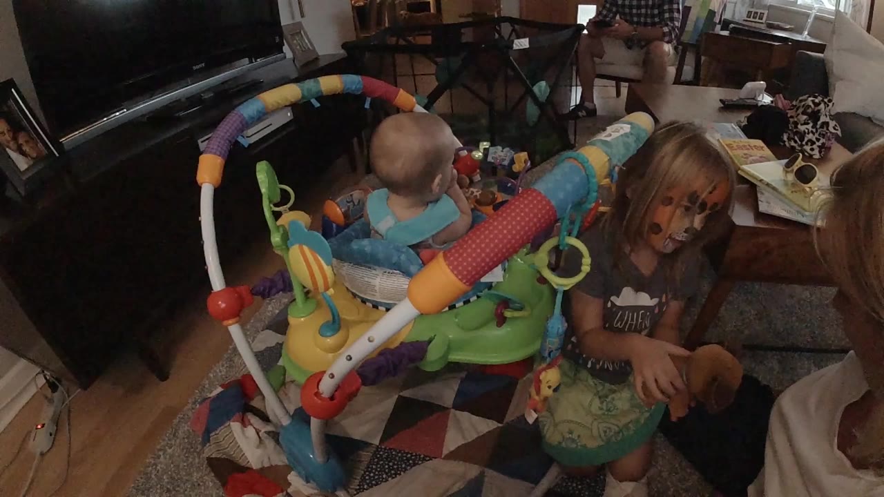 In a Colorado home interior, a baby sits in a colorful activity jumper while a blonde girl with face paint stands nearby with toys around the room.