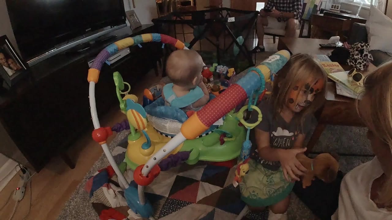 A blonde girl with painted cheeks leans near the baby in the activity jumper, with the black TV and scattered home items behind them.