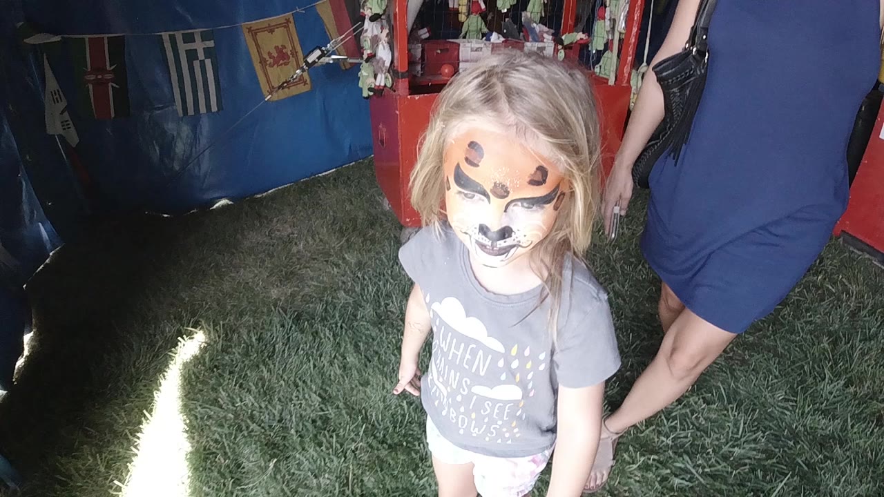 A young blonde girl with animal-style face paint stands on grass near the opening of a blue tent in Colorado, with an adult partly visible beside her.