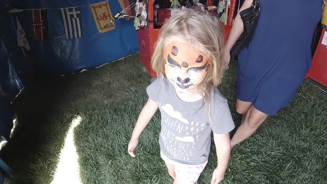 The young girl with face paint begins moving toward the brighter tent exit, with grass underfoot and the adult still partly cropped at the right edge.