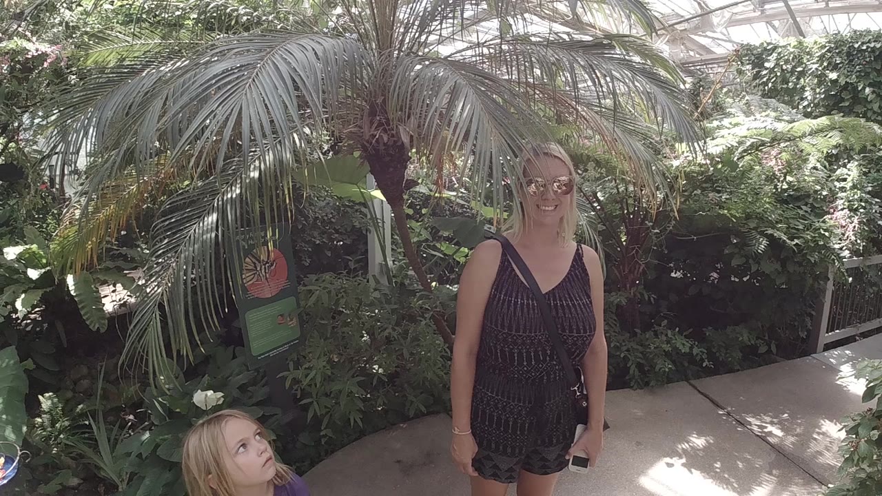 In Colorado, the woman smiles under the palm canopy while the child's face remains partly visible near the lower-left edge and a plant sign sits behind the foliage.