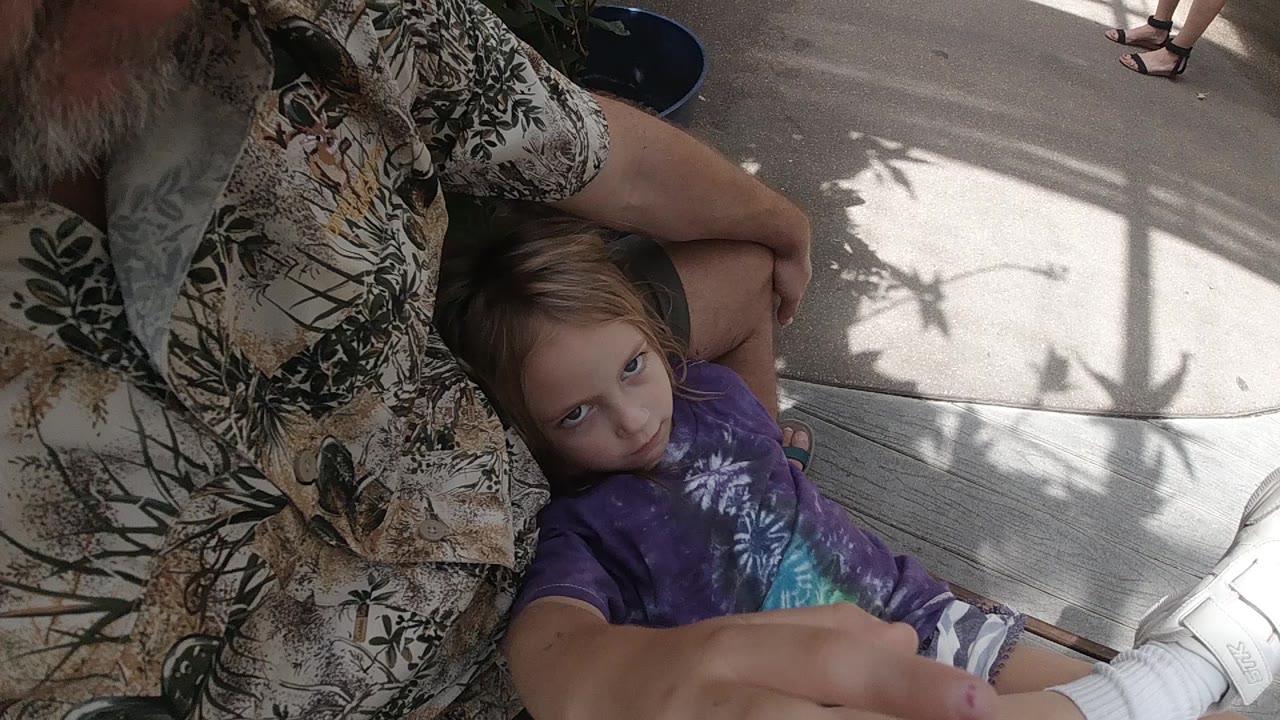 In Colorado, a young blonde girl in a purple shirt sits on outdoor decking beside a patterned-shirt adult and looks up before reaching fully forward.