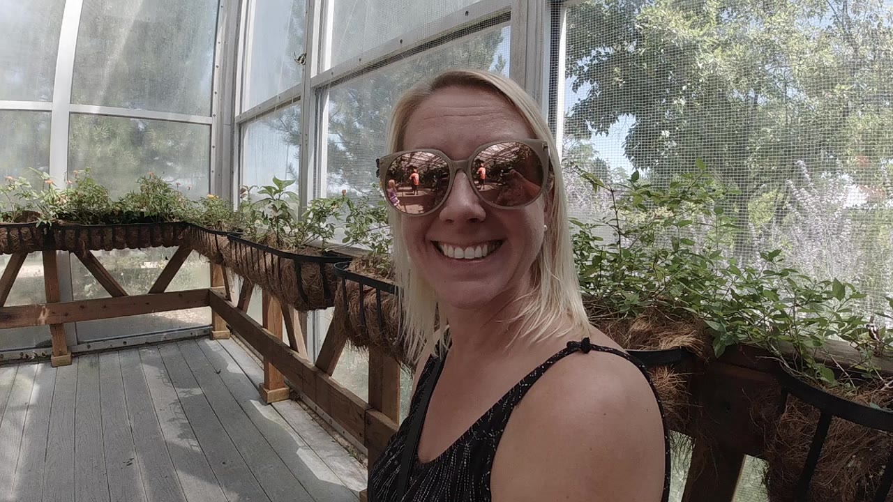 At the start of the Denver clip, a blonde woman in reflective sunglasses smiles at the camera inside a bright greenhouse-like garden space with plant benches behind her.