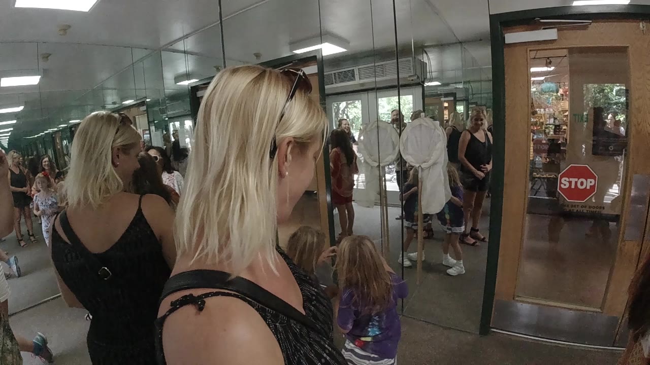 The blonde woman is viewed mostly from behind in the Colorado interior, with children seated and standing along the glass wall and a door marked by a red stop sign.