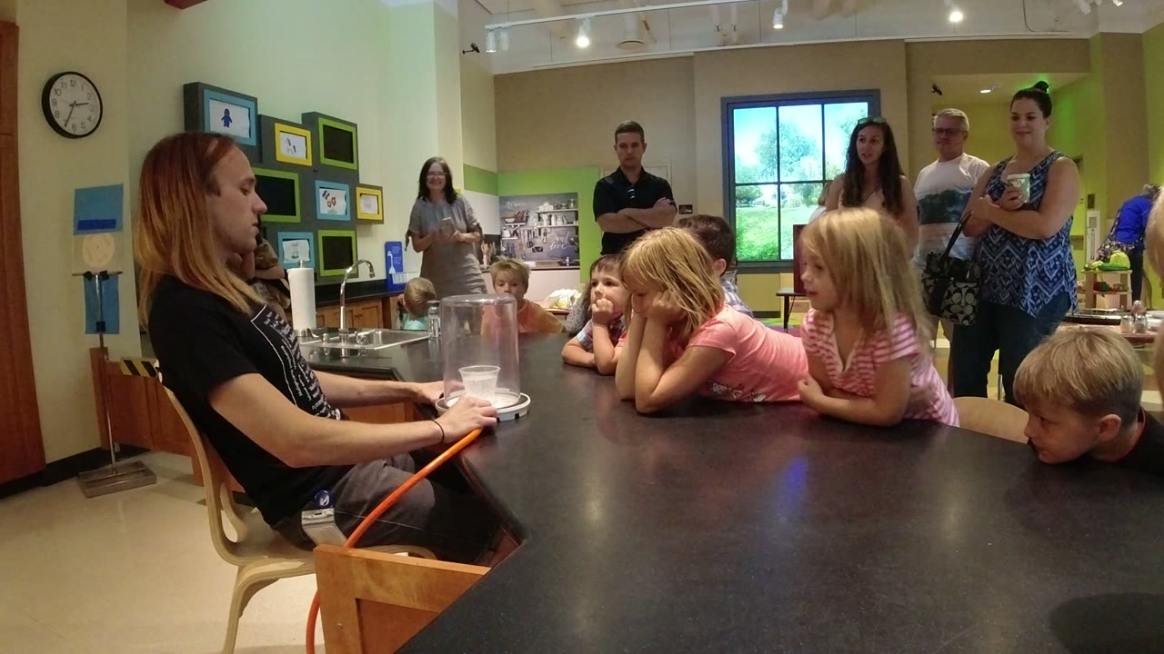 Children in Colorado rest their arms on the table and look toward the demonstration equipment while adults observe near the rear window and wall displays.