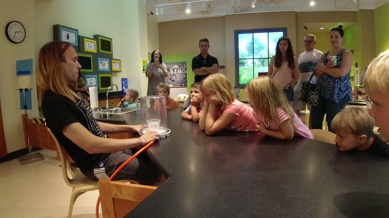 Presenter sits beside children gathered around a science table.