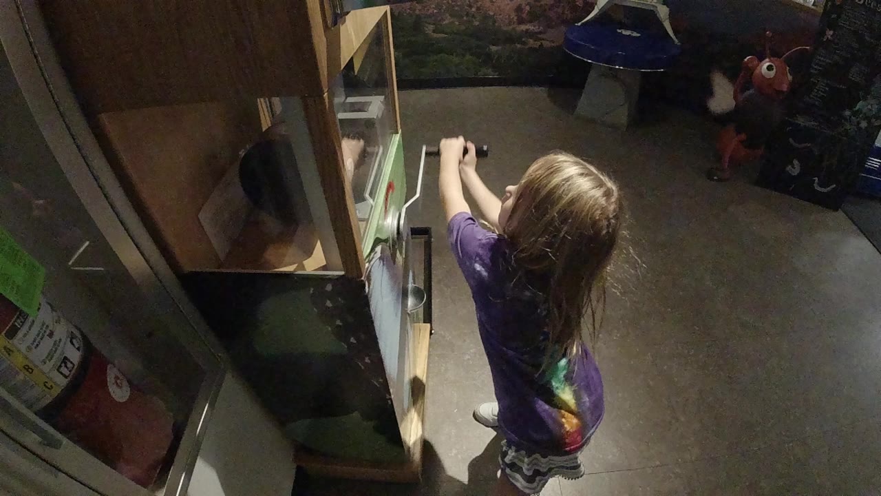 A young girl in a purple shirt stands below an open refrigerator, viewed from above in a dim kitchen.