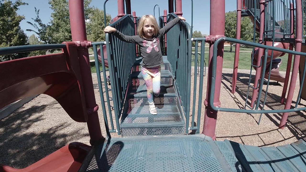A little girl stands near the far end of a teal metal playground bridge in a Colorado park, with red posts and a slide edge around her.