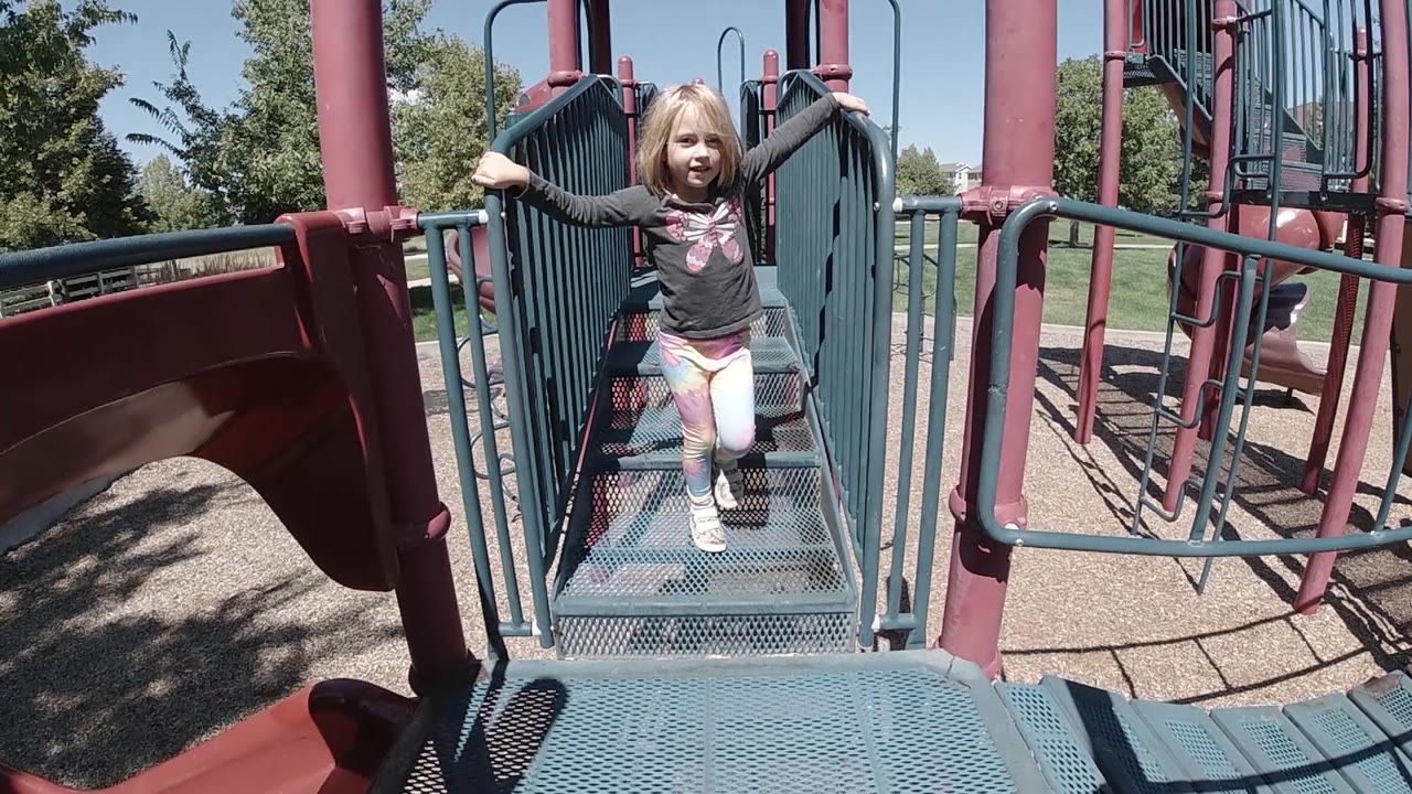 The girl moves closer along the teal bridge, with one hand on each railing and the Colorado park grass visible through the bars.