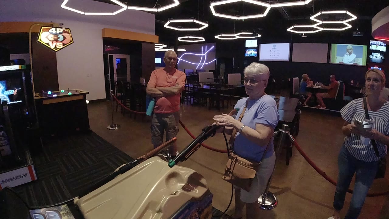 The senior woman keeps both hands on the mounted rifle in the Denver indoor entertainment space as a man in a coral shirt watches from behind.