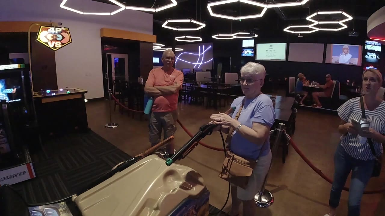 The senior player looks down toward the mounted rifle while a striped-shirt woman stands partly in frame at right and arcade screens glow in the background.