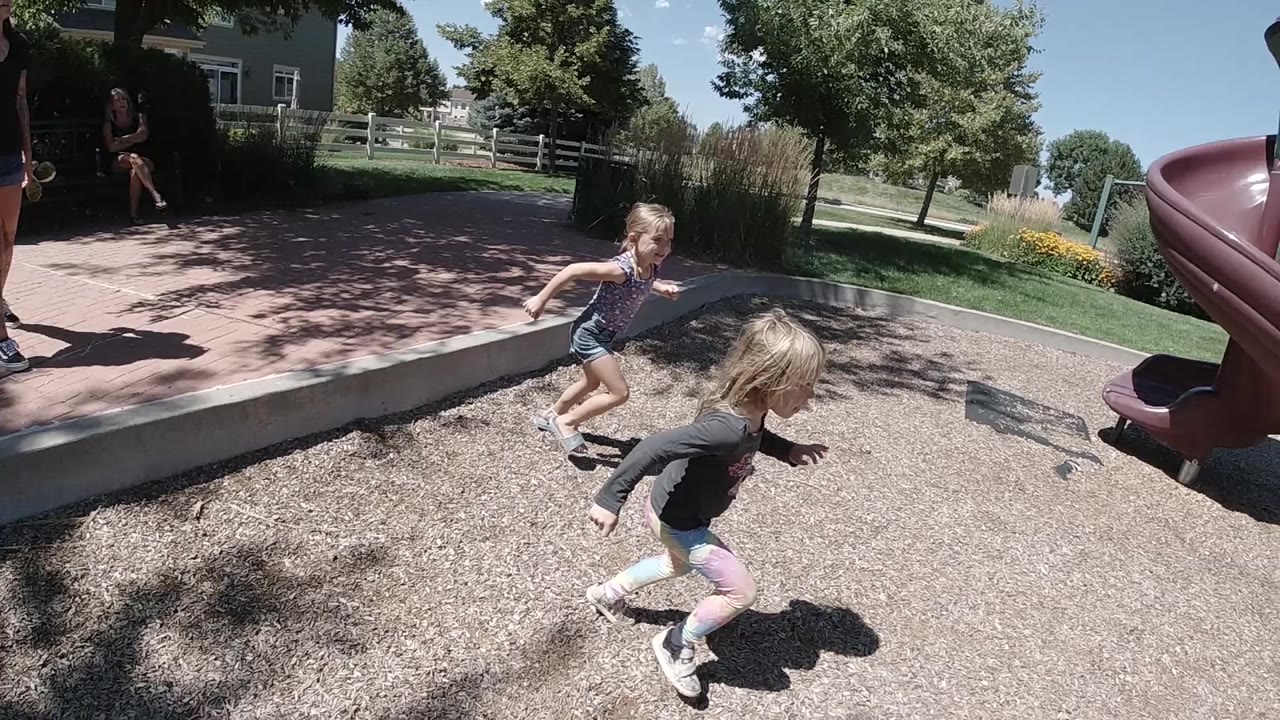 Two young girls run across playground mulch near a red spiral slide.