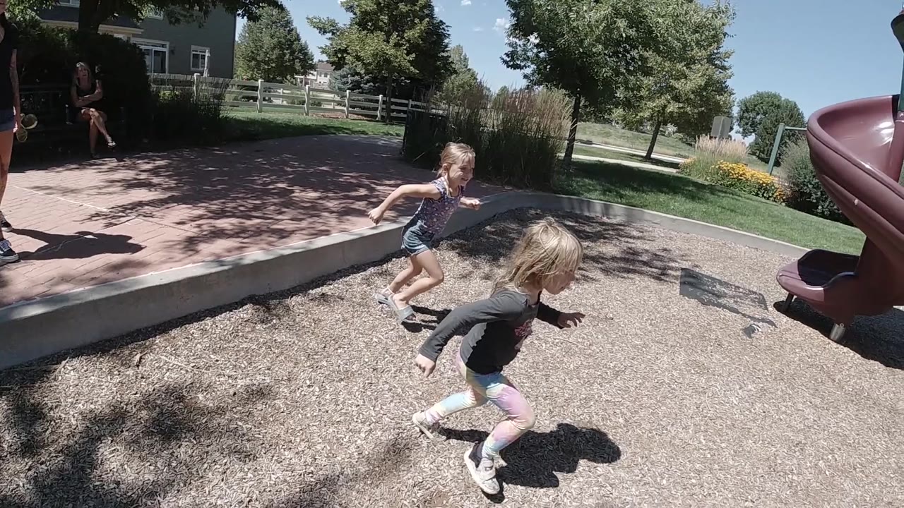 The girls run across the mulch toward the play structure, with the red slide on the right and a sunny park background behind them.
