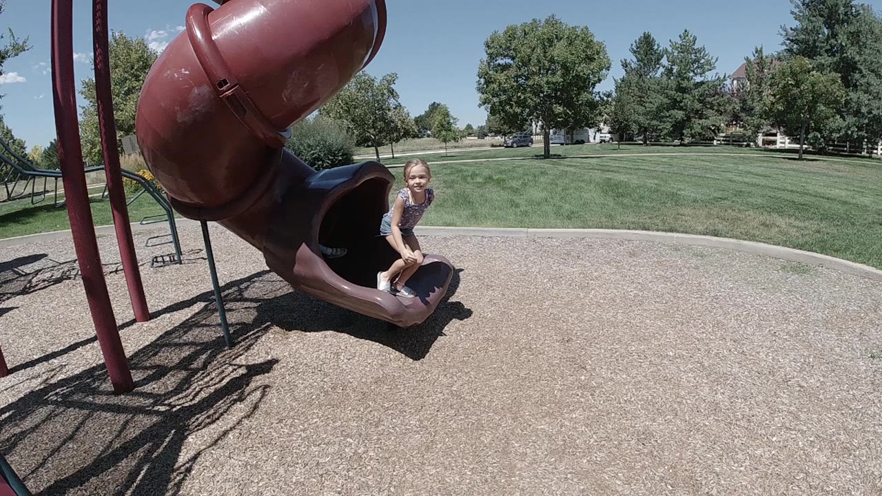 A young girl sits at the bottom opening of a maroon tubular playground slide in Denver, with grass, trees, and homes beyond the play surface.