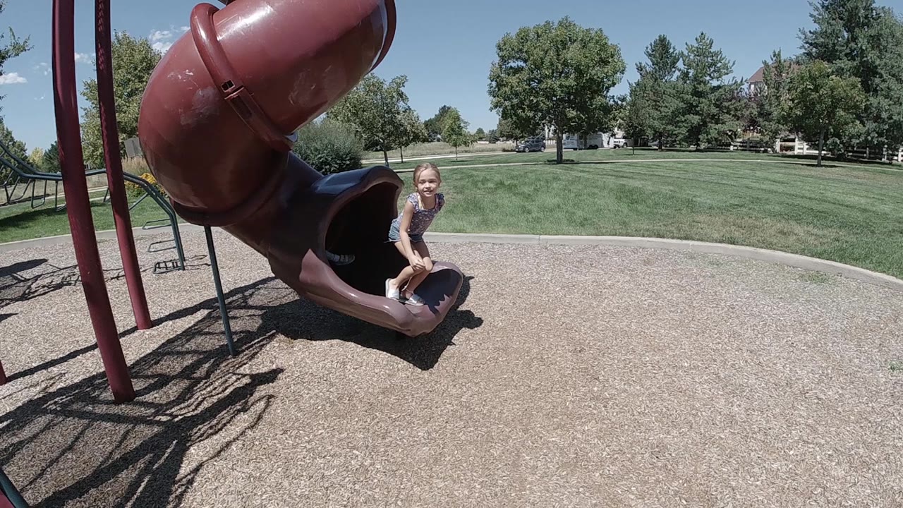 The child stays seated near the slide mouth in Denver while the wide view shows tan playground surfacing and a broad green lawn in bright sun.