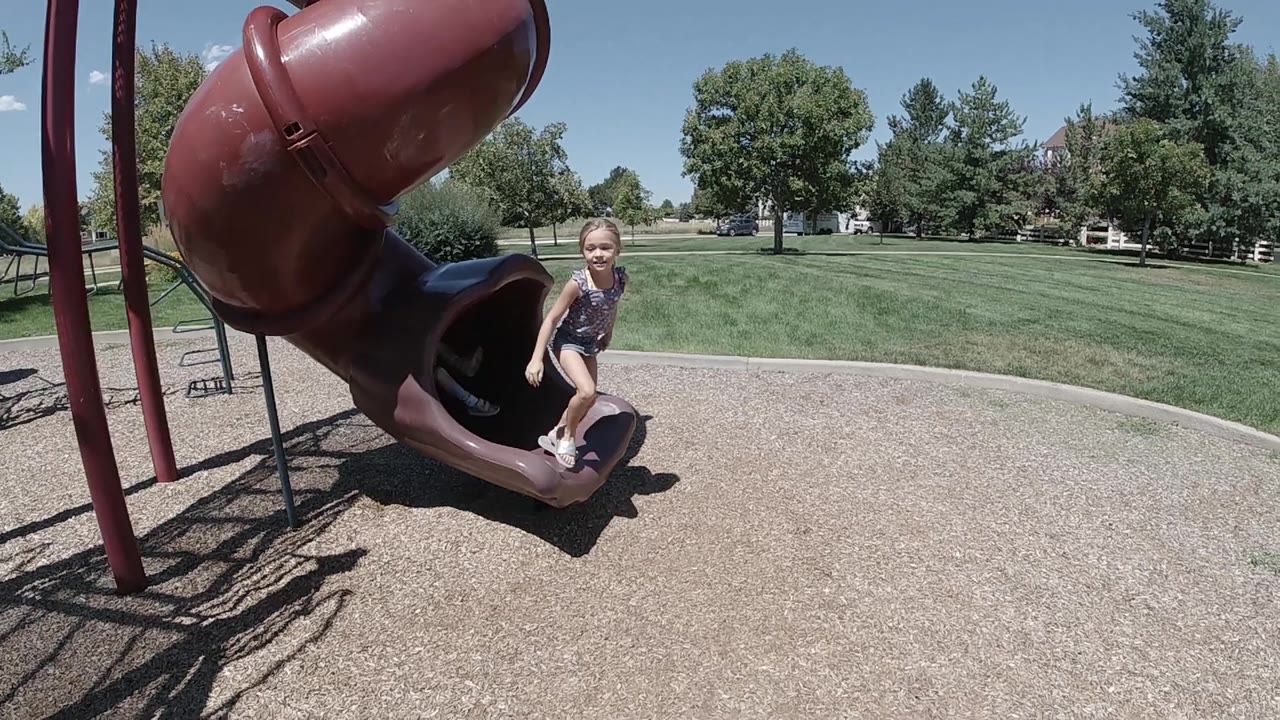 A young girl starts leaving the maroon tube slide in Denver, with one hand braced near the slide edge and open lawn stretching behind her.