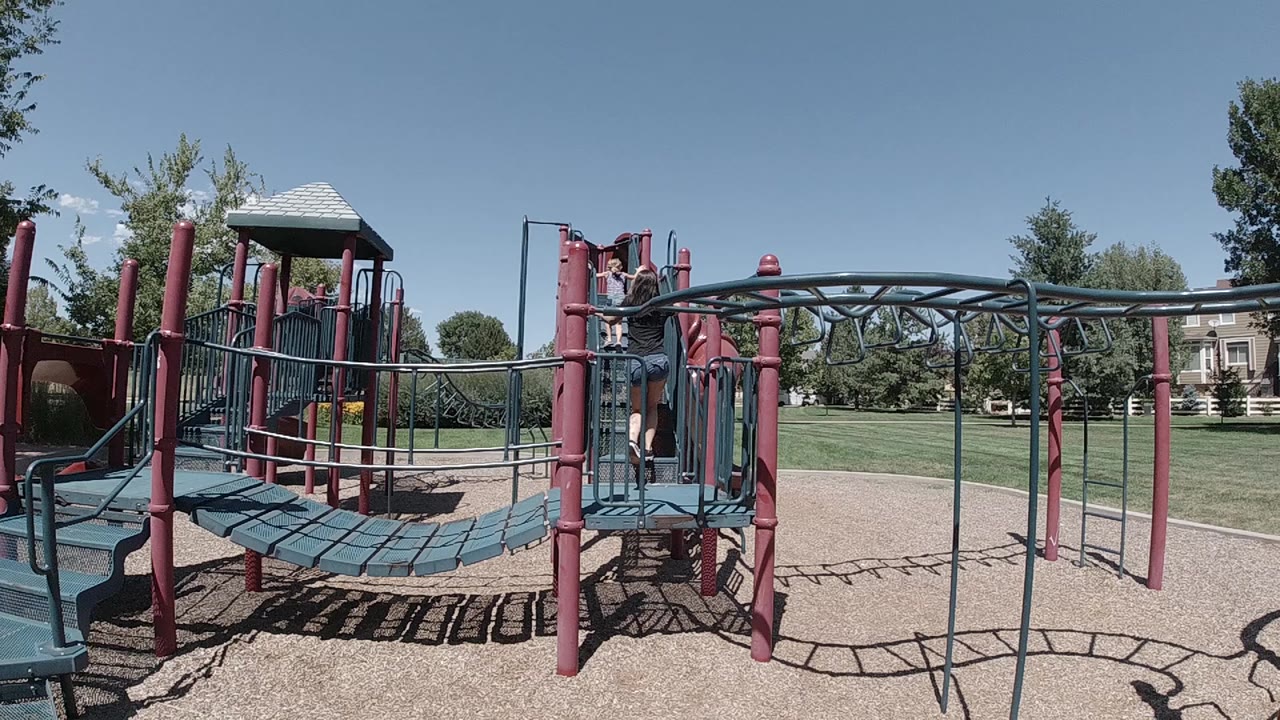 Wide view of a red and teal playground in Colorado with a child near the left platform, grass, trees, and clear sky in the background.