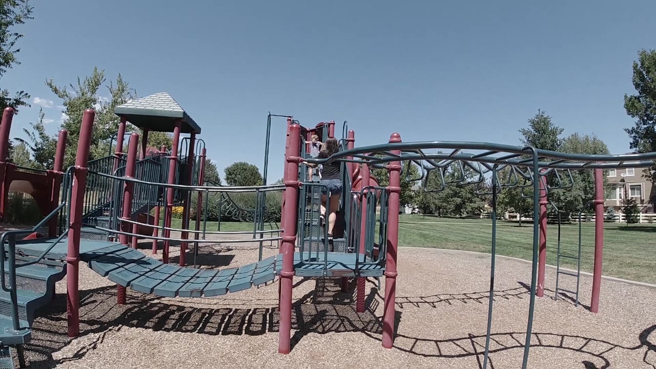 A child is visible on the raised walkway of the Colorado playground, with curved teal monkey bars spanning the right side of the frame.