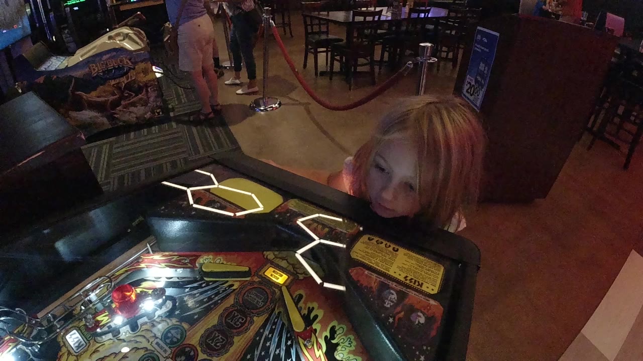 Young blonde girl peers over a lit pinball machine in an arcade.