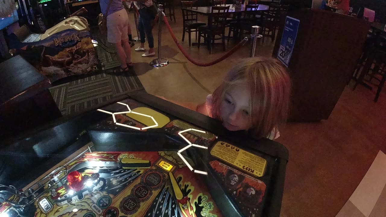 At 0:01 in Denver, the girl's face sits low beside the pinball cabinet as bright playfield lights reflect across the glass.